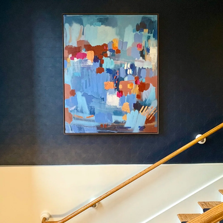 An abstract framed painting with blue, orange, brown, and white brushstrokes hanging on a dark navy wall above a staircase with a wooden handrail.